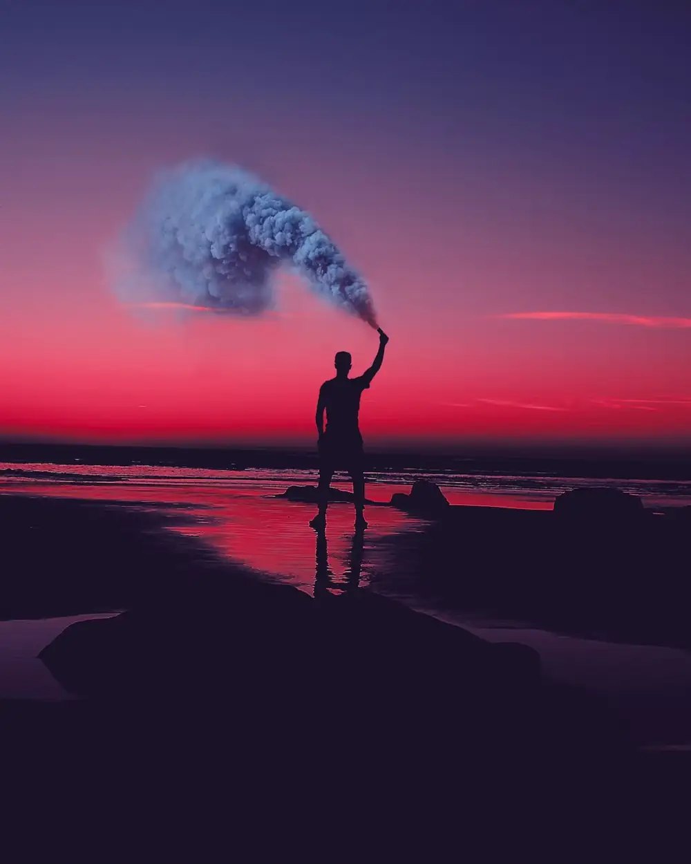 A figure on the shore raising a smoke flare into a crimson sunset sky